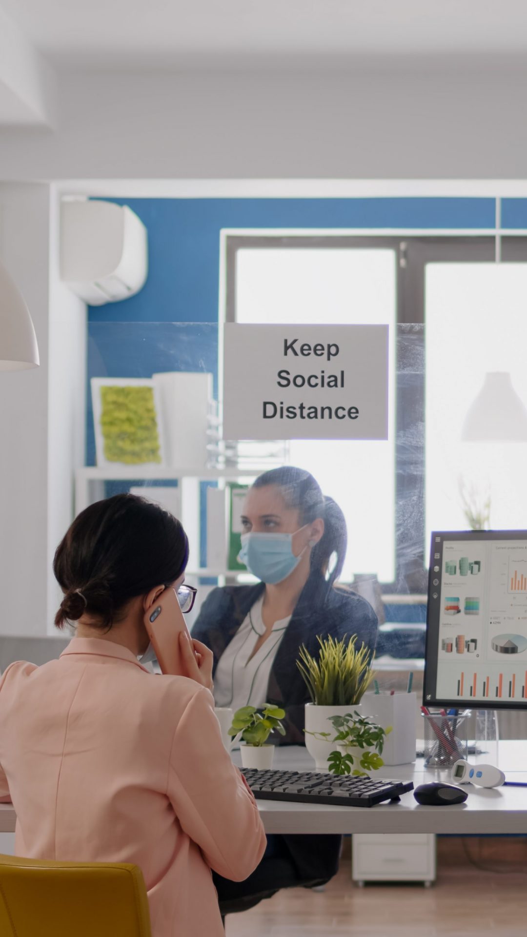 business-woman-with-medical-face-mask-talking-smartphone-with-digital marketing expert-while-her-coworkers