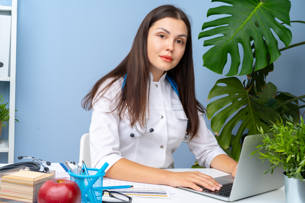 Woman doctor portrait at her office desk, office interior using laptop and updating roaster on health care website