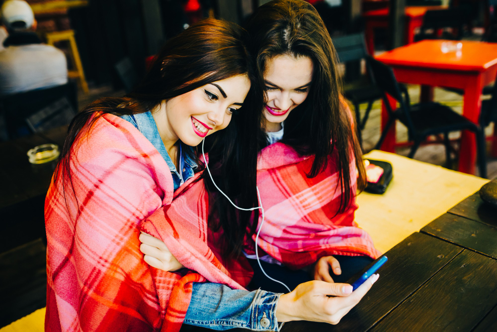 Two young and beautiful girl sitting at the table listening to music on mobile design website